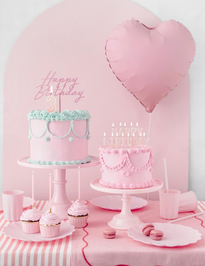 May include: Two pink cakes with pink candles on cake stands. The cakes are decorated with pink frosting and white accents. A pink heart-shaped balloon is in the background. The text "Happy Birthday" is on a pink cake topper.
