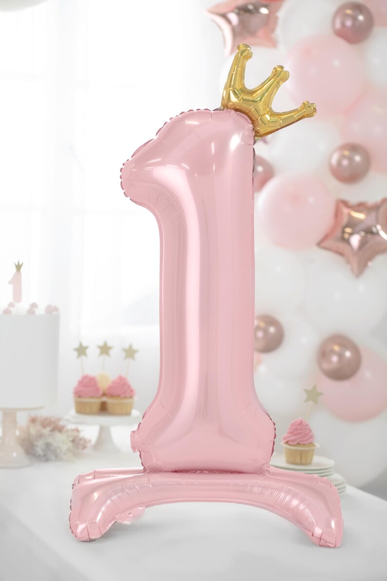 May include: A pink number one balloon with a gold crown on top. The balloon is standing on a pink base.