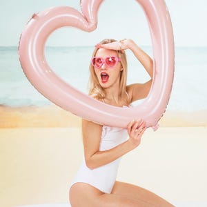 Woman holding a large light pink heart shaped balloon frame on the beach, summer party decoration and photo prop