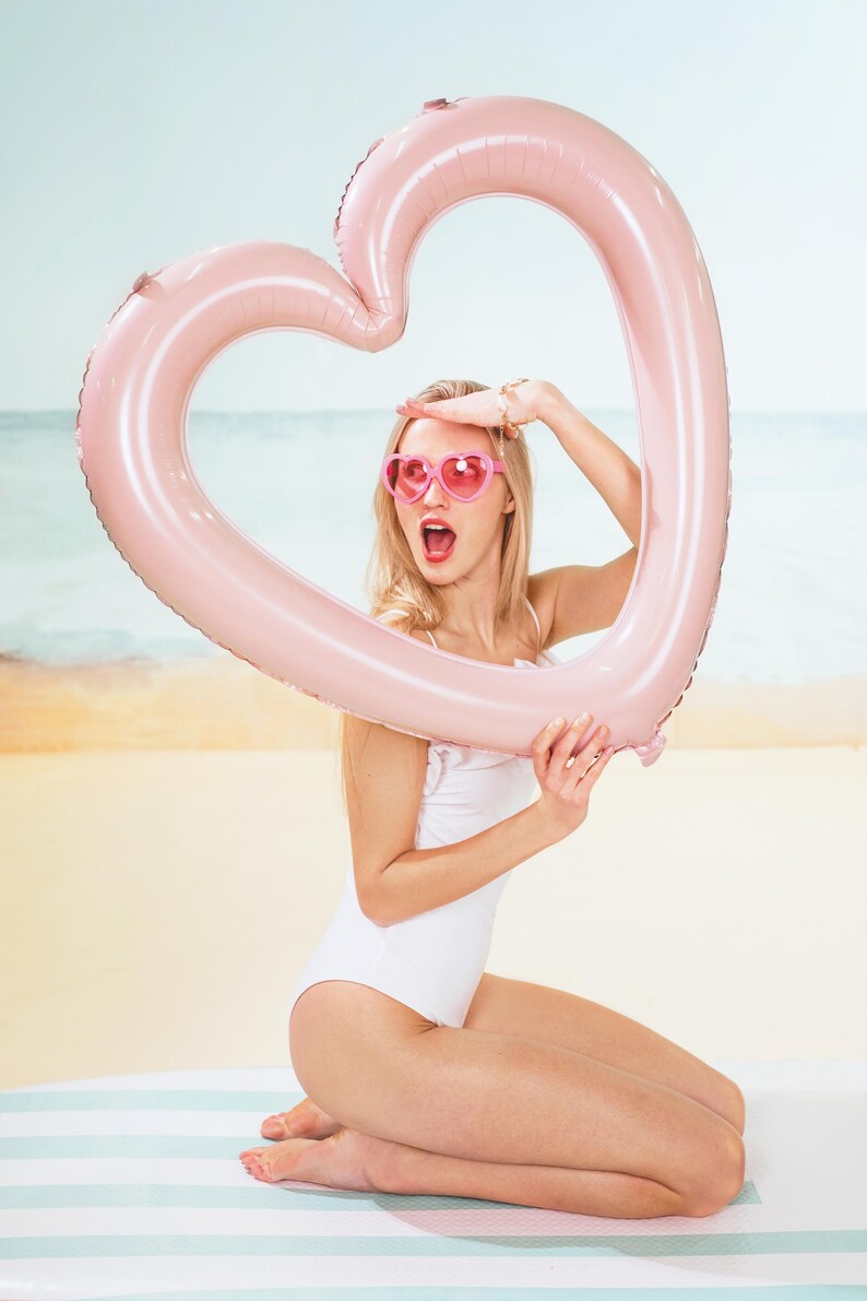 Woman holding a large light pink heart shaped balloon frame on the beach, summer party decoration and photo prop