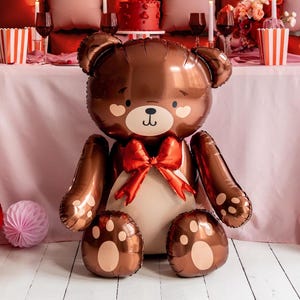 Pu&ograve; includere: Un grande palloncino a forma di orsacchiotto marrone con un fiocco rosso e un viso carino. Il palloncino ha un naso e occhi a forma di cuore.