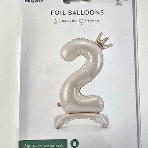 Peut inclure: Un ballon en feuille d'aluminium blanc en forme de chiffre 2 avec une couronne dor&eacute;e sur le dessus. Le ballon mesure 103 cm de haut et 84 cm de large. Le ballon est sur un fond blanc avec le texte "FOIL BALLOONS" et "PartyDeco" imprim&eacute; sur le fond.