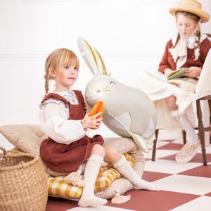 May include: Two young girls playing with a large, gray, rabbit-shaped balloon. One girl is holding the balloon while the other girl is sitting on a checkered floor with a book.