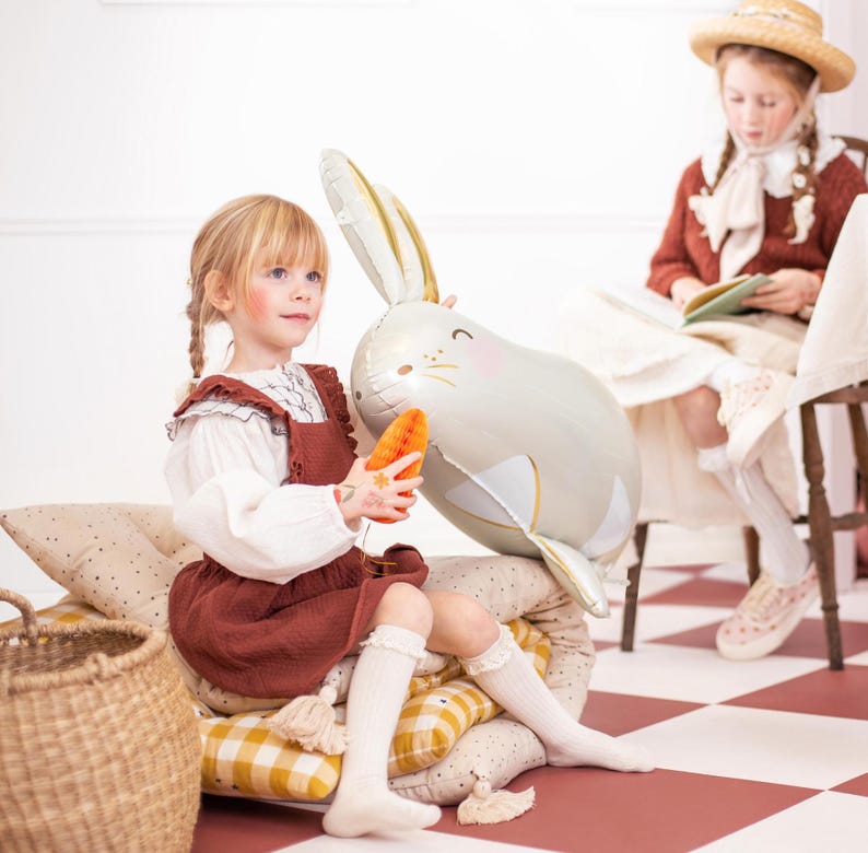 May include: Two young girls playing with a large, gray, rabbit-shaped balloon. One girl is holding the balloon while the other girl is sitting on a checkered floor with a book.