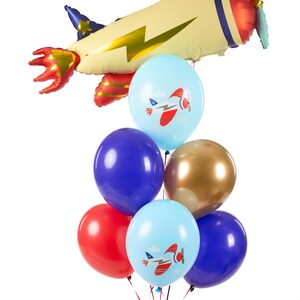 K&ouml;nnte beinhalten: Ein B&uuml;ndel bunter Ballons mit Flugzeugmotiven, darunter ein gro&szlig;er Flugzeugballon in Gelb, Rot und Gold. Die Ballons werden von einer Hand gehalten, die ein rot-wei&szlig; gestreiftes Hemd tr&auml;gt.