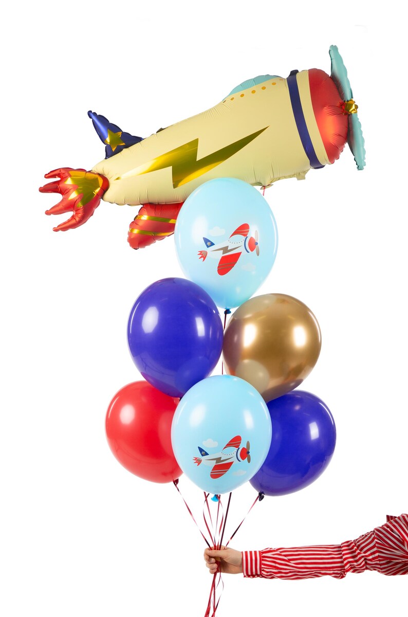 K&ouml;nnte beinhalten: Ein B&uuml;ndel bunter Ballons mit Flugzeugmotiven, darunter ein gro&szlig;er Flugzeugballon in Gelb, Rot und Gold. Die Ballons werden von einer Hand gehalten, die ein rot-wei&szlig; gestreiftes Hemd tr&auml;gt.