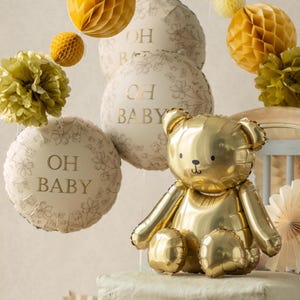 Puede incluir: Un globo de oso dorado se encuentra entre globos color crema con la inscripci&oacute;n "OH BABY" en dorado. Bolas decorativas de panal de papel y detalles florales en amarillo y verde completan el tema del baby shower.