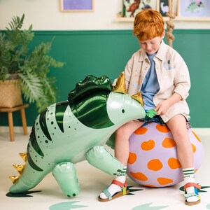 Puede incluir: Un ni&ntilde;o se sienta en una pelota grande de lunares naranja y morado, sosteniendo un dinosaurio inflable verde y blanco. El dinosaurio tiene una espalda verde con p&uacute;as y una cara blanca.