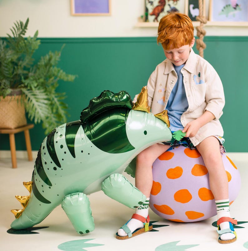 Puede incluir: Un ni&ntilde;o se sienta en una pelota grande de lunares naranja y morado, sosteniendo un dinosaurio inflable verde y blanco. El dinosaurio tiene una espalda verde con p&uacute;as y una cara blanca.