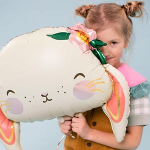 May include: A white bunny balloon with pink ears and a pink flower on its head. The balloon has a cute cartoon face with black eyes and a pink nose. A child is holding the balloon in front of their face.