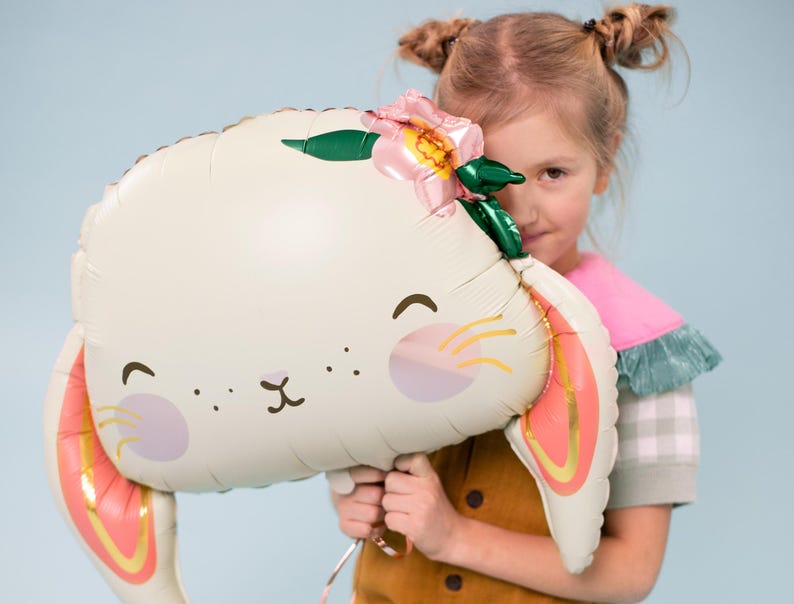 May include: A white bunny balloon with pink ears and a pink flower on its head. The balloon has a cute cartoon face with black eyes and a pink nose. A child is holding the balloon in front of their face.