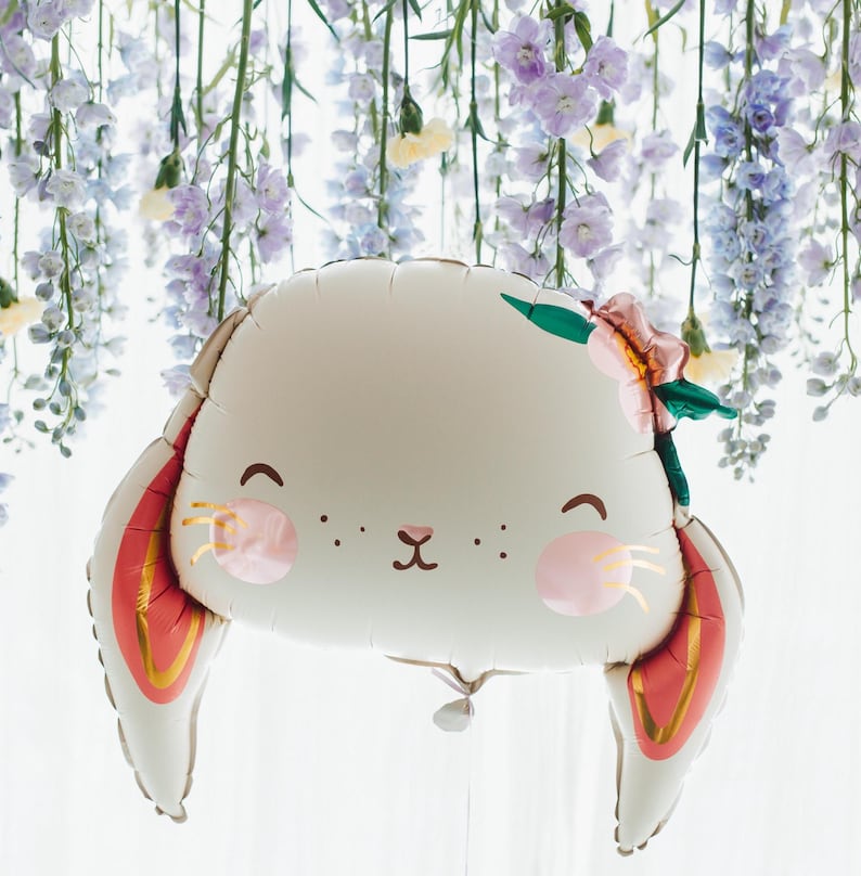 May include: A white balloon shaped like a bunny with red and gold ears. The balloon has a cute face with pink cheeks and a small black nose. The balloon is decorated with a green leaf and a pink flower.