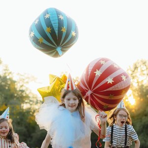 K&ouml;nnte beinhalten: Kinder feiern im Freien mit Partyballons. Zwei gro&szlig;e, quadratische Ballons mit Stern- und Streifenmustern in Rot, Blau und Gold. Die Kinder tragen Partyh&uuml;te und l&auml;cheln, genie&szlig;en die Feierlichkeiten.