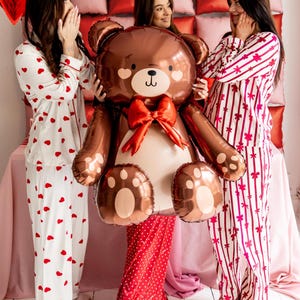 Pu&ograve; includere: Tre persone in pigiama posano con un grande palloncino a forma di orsacchiotto di peluche marrone. Il palloncino ha un fiocco rosso e un viso carino. I pigiami sono bianchi con cuori rossi, rossi con pois bianchi e bianchi con strisce rosa e rosse.