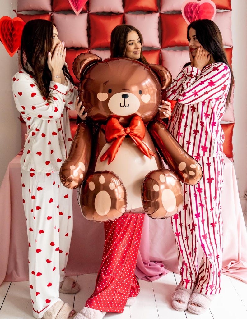 Pu&ograve; includere: Tre persone in pigiama posano con un grande palloncino a forma di orsacchiotto di peluche marrone. Il palloncino ha un fiocco rosso e un viso carino. I pigiami sono bianchi con cuori rossi, rossi con pois bianchi e bianchi con strisce rosa e rosse.