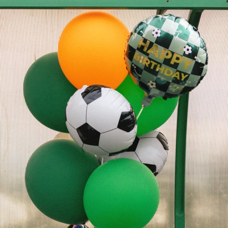 May include: A festive balloon bouquet featuring green, orange, and soccer ball-themed balloons. One balloon reads "HAPPY BIRTHDAY" in gold lettering. The balloons are arranged together, creating a celebratory display.