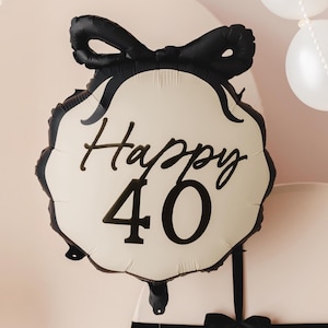A festive scene featuring a cream-colored and black balloon with a bow design and the text "Happy 40" in elegant black lettering. The background includes white balloons and pearl-like garlands, along with large, round, cream-colored props tied with black ribbon, creating a sophisticated 40th celebration setup.
