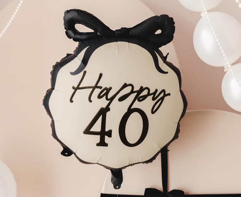 A festive scene featuring a cream-colored and black balloon with a bow design and the text "Happy 40" in elegant black lettering. The background includes white balloons and pearl-like garlands, along with large, round, cream-colored props tied with black ribbon, creating a sophisticated 40th celebration setup.