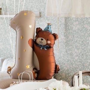 May include: A brown bear balloon wearing a blue party hat is hugging a large beige number one balloon with gold stars. The balloons are on a white surface.