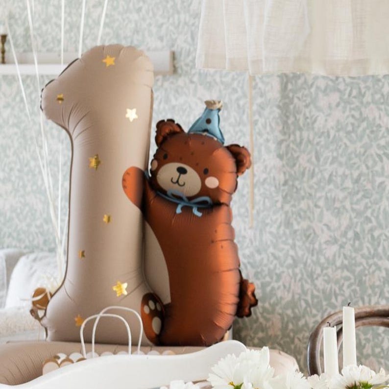 May include: A brown bear balloon wearing a blue party hat is hugging a large beige number one balloon with gold stars. The balloons are on a white surface.