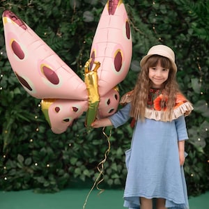 May include: A young girl wearing a blue dress and a straw hat holds a pink butterfly balloon with gold accents. The balloon is tied to a gold tassel. The girl is standing in front of a green leafy background.