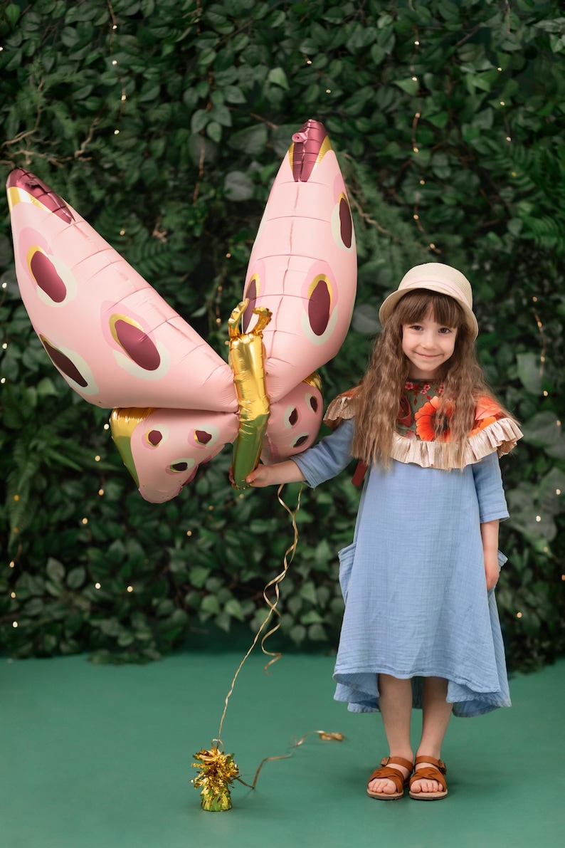 May include: A young girl wearing a blue dress and a straw hat holds a pink butterfly balloon with gold accents. The balloon is tied to a gold tassel. The girl is standing in front of a green leafy background.