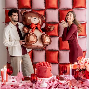 Pu&ograve; includere: Uno sfondo di palloncini rossi e rosa con un palloncino a forma di orsacchiotto marrone, una torta rossa con glassa bianca e due persone sorridenti.