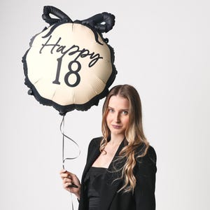 May include: A woman holds a black and cream balloon with the text "Happy 18" in black script. She is wearing a black blazer and has long brown hair.