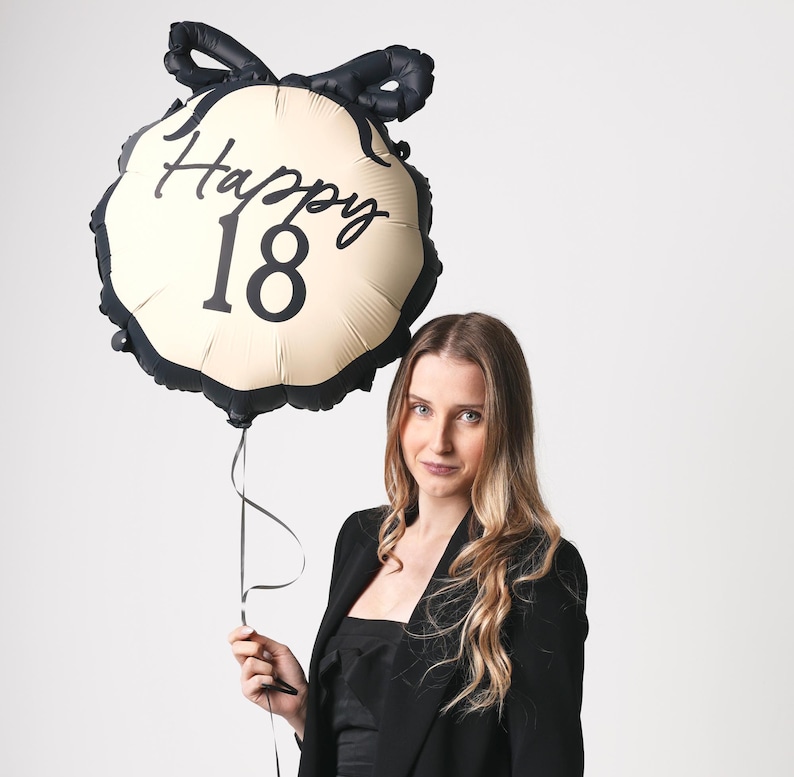 May include: A woman holds a black and cream balloon with the text "Happy 18" in black script. She is wearing a black blazer and has long brown hair.