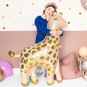 May include: A young girl hugs a large gold and beige giraffe balloon. The balloon has a spotted pattern and a friendly face with eyelashes.