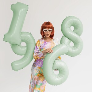 Pu&ograve; includere: Una donna con un vestito floreale e occhiali da sole tiene un set di palloncini di alluminio verde a forma dei numeri 4 e 8.