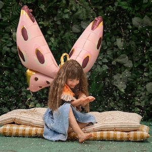 May include: A young girl wearing a blue dress and a pink butterfly costume sits on a striped cushion. She is surrounded by a green leafy background and sparkling glitter.