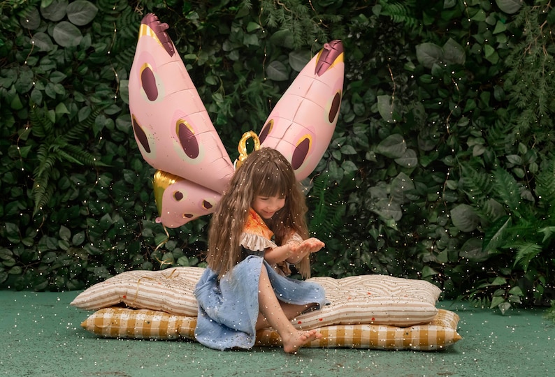 May include: A young girl wearing a blue dress and a pink butterfly costume sits on a striped cushion. She is surrounded by a green leafy background and sparkling glitter.
