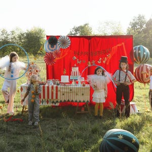 May include: A festive outdoor circus-themed birthday party scene. Children in costumes stand in a grassy area with a decorated table, red backdrop, and balloons. The scene includes a cake, decorations, and a "Happy Birthday" banner.