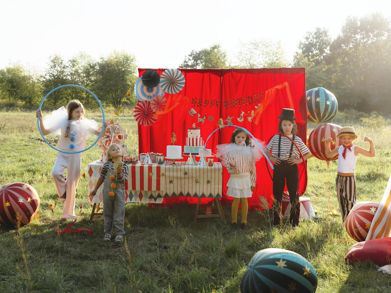 May include: A festive outdoor circus-themed birthday party scene. Children in costumes stand in a grassy area with a decorated table, red backdrop, and balloons. The scene includes a cake, decorations, and a "Happy Birthday" banner.