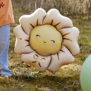 Puede incluir: Un gran globo de color crema en forma de flor con una cara de sol amarilla y un lazo dorado. El globo tiene una expresi&oacute;n feliz con los ojos cerrados y las mejillas sonrosadas.