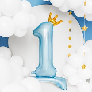 K&ouml;nnte beinhalten: Ein blauer Ballon in Form der Zahl eins mit einer goldenen Krone oben drauf. Der Ballon ist umgeben von wei&szlig;en Ballons und einer goldenen Sternen-Girlande.