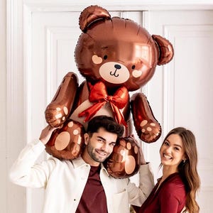 Pu&ograve; includere: Un grande palloncino a forma di orsacchiotto marrone con un fiocco rosso, tenuto da due persone. Il palloncino ha un viso da cartone animato carino con occhi neri e un naso marrone.
