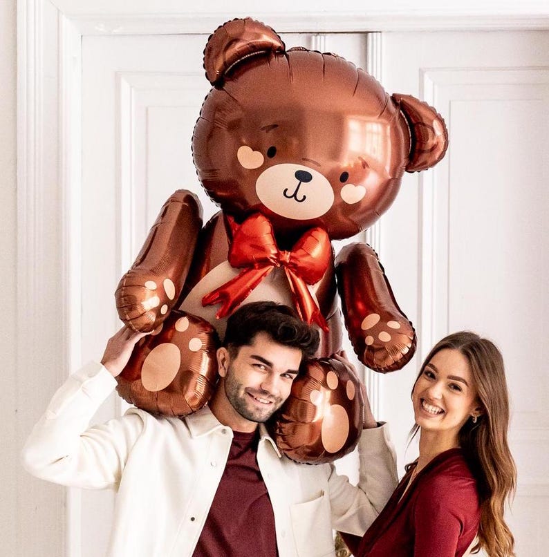 Pu&ograve; includere: Un grande palloncino a forma di orsacchiotto marrone con un fiocco rosso, tenuto da due persone. Il palloncino ha un viso da cartone animato carino con occhi neri e un naso marrone.