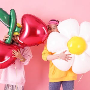 May include: Two girls holding large foil balloons. One balloon is a red cherry and the other is a white daisy. The girl holding the cherry balloon is wearing a pink hoodie and the girl holding the daisy balloon is wearing a yellow hoodie.