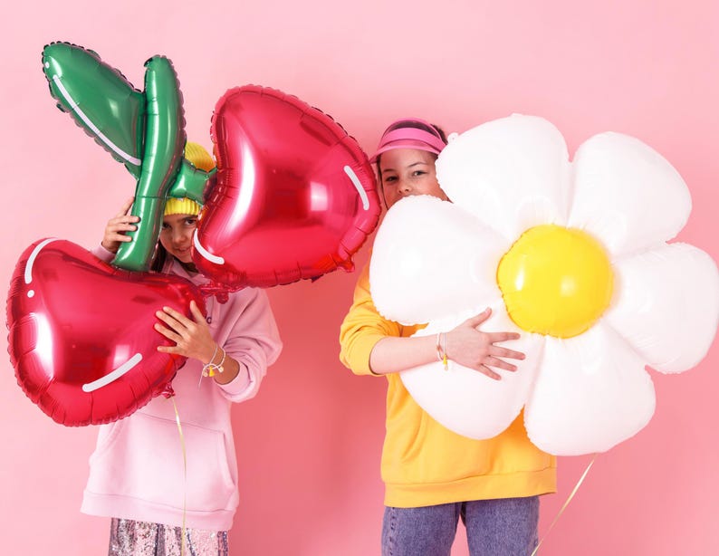 May include: Two girls holding large foil balloons. One balloon is a red cherry and the other is a white daisy. The girl holding the cherry balloon is wearing a pink hoodie and the girl holding the daisy balloon is wearing a yellow hoodie.
