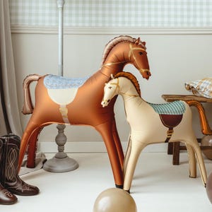 Puede incluir: Dos juguetes inflables de caballos, uno grande marr&oacute;n con una silla de montar azul claro y otro m&aacute;s peque&ntilde;o crema con una silla de montar verde y marr&oacute;n. Est&aacute;n sobre un suelo blanco, con una pared a cuadros azul claro y blanco al fondo.