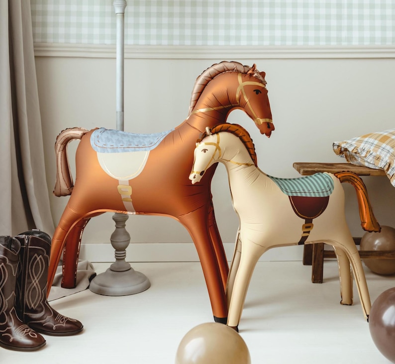 Puede incluir: Dos juguetes inflables de caballos, uno grande marr&oacute;n con una silla de montar azul claro y otro m&aacute;s peque&ntilde;o crema con una silla de montar verde y marr&oacute;n. Est&aacute;n sobre un suelo blanco, con una pared a cuadros azul claro y blanco al fondo.