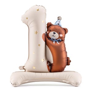 A standing balloon beige number 1-shaped with to the side is a brown bear-shaped wearing a blue party hat and a blue bow. The entire arrangement is set on a beige inflatable base.