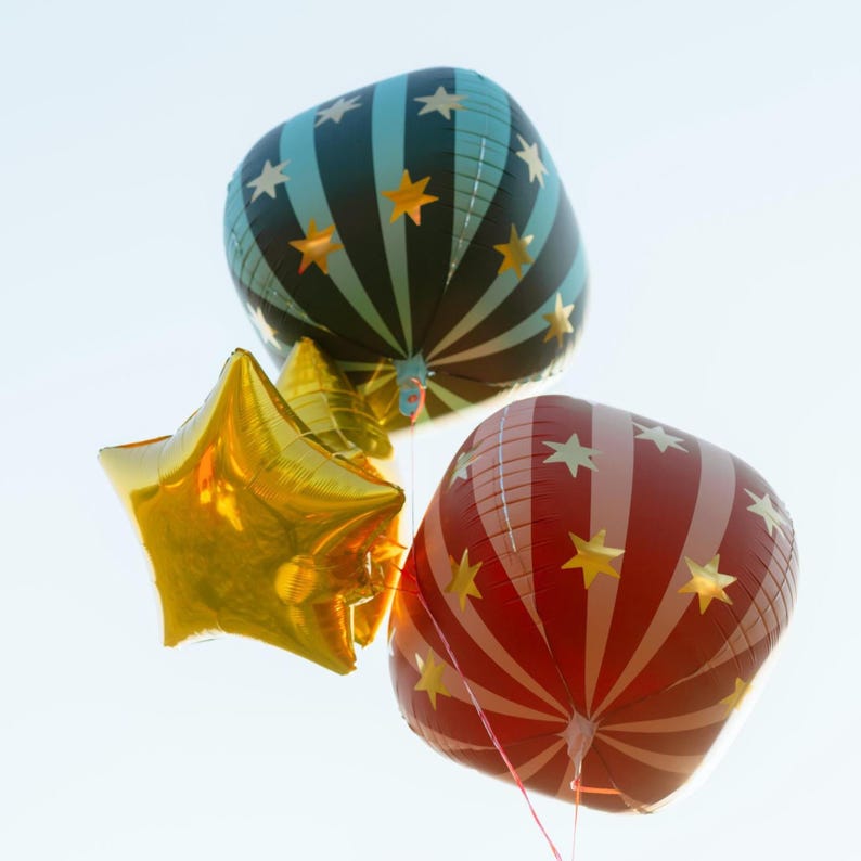 May include: Three helium balloons against a clear sky. One is a gold star shape, and the other two are square-shaped with star and stripe patterns. One square balloon is black and teal, and the other is red and white.