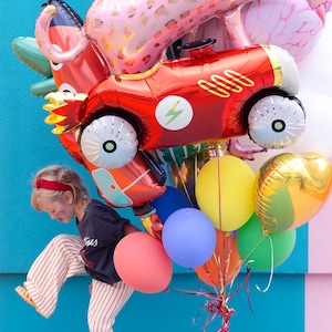 May include: A child holds a bunch of colorful balloons, including a red race car balloon, a pink leopard balloon, and a yellow balloon. The balloons are tied together with red and gold ribbon.