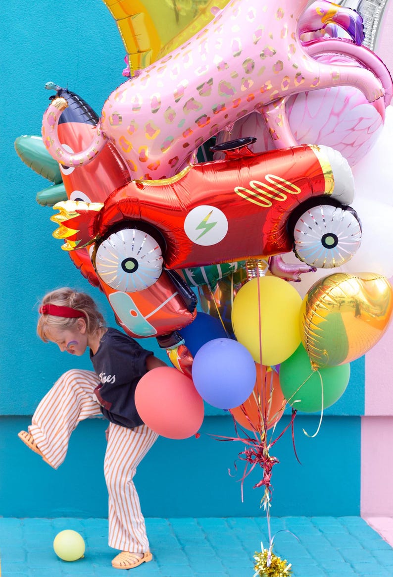 May include: A child holds a bunch of colorful balloons, including a red race car balloon, a pink leopard balloon, and a yellow balloon. The balloons are tied together with red and gold ribbon.