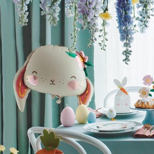May include: A white bunny balloon with pink cheeks and a flower crown, along with pastel colored eggs and a white bunny gift bag, set on a light blue table with a white tablecloth. The table is decorated with a floral garland.