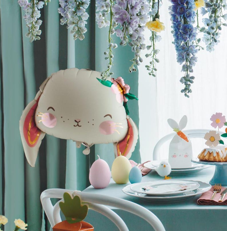 May include: A white bunny balloon with pink cheeks and a flower crown, along with pastel colored eggs and a white bunny gift bag, set on a light blue table with a white tablecloth. The table is decorated with a floral garland.