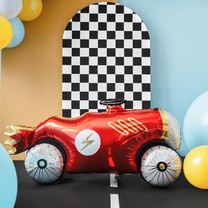 May include: A red race car shaped foil balloon with a lightning bolt design and gold accents. The balloon is in front of a black and white checkered background.